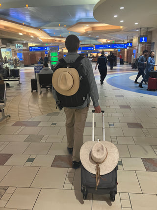 Charlie walking through airport with his two hats clipped to his bags. one is clipped to his rolling luggage and the other to his backpack to keep it safe.